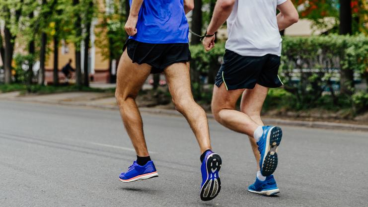 Zwei Männer joggen auf einer Straße, beide in Sportkleidung und Laufschuhen, umgeben von grünen Bäumen.