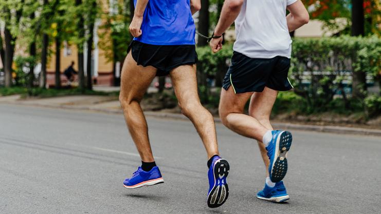 Zwei Männer joggen auf einer Straße, beide in Sportkleidung und Laufschuhen, umgeben von grünen Bäumen.