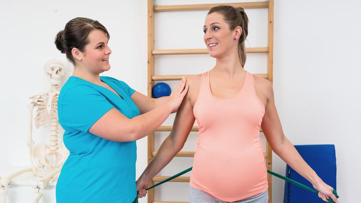 Physiotherapeutin unterstützt schwangere Frau mit einem grünen Theraband in einem modernen Behandlungsraum.