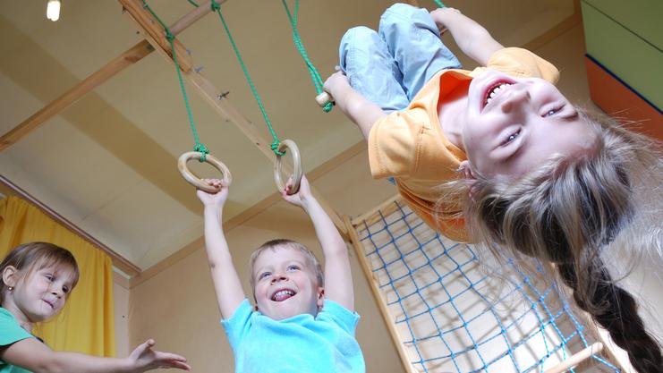 Drei Kinder spielen in einem Raum: Ein Junge hängt an Ringen, während zwei andere lachen und zuschauen.