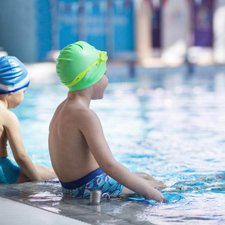 Zwei Kinder in Schwimmkappen sitzen am Beckenrand eines Hallenbades und beobachten das Wasser.