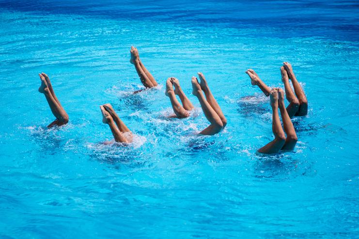 Synchronschwimmer führen eine Choreografie mit gestreckten Beinen im blauen Wasser aus. Spritzwasser ist sichtbar.