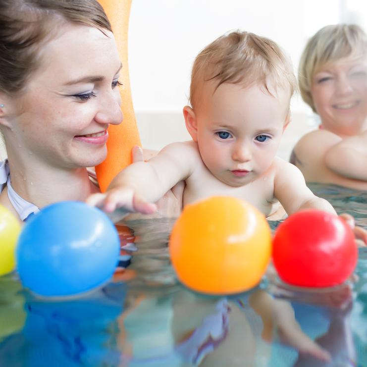 Zwei Erwachsene und zwei Kleinkinder spielen im Wasser mit bunten Plastikbällen in einem Schwimmbad.