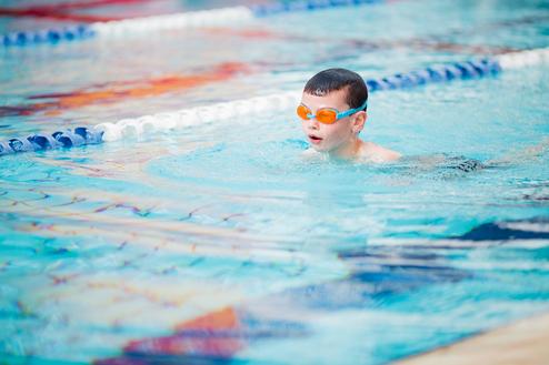 Junge mit orangenen Schwimmbrille schwimmt im Pool, Wasser spritzt um ihn herum, blaue Bahnen sind im Hintergrund sichtbar.