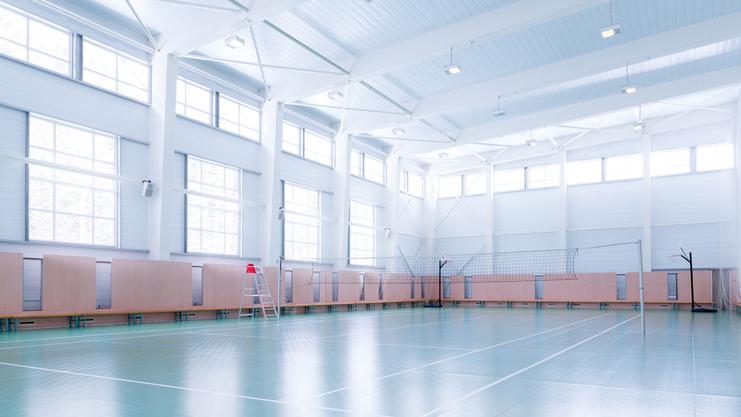 Leere Sporthalle mit hellem Boden, Fensterfront und Volleyballnetzen an der Seite. Eine Leiter steht neben dem Netz.